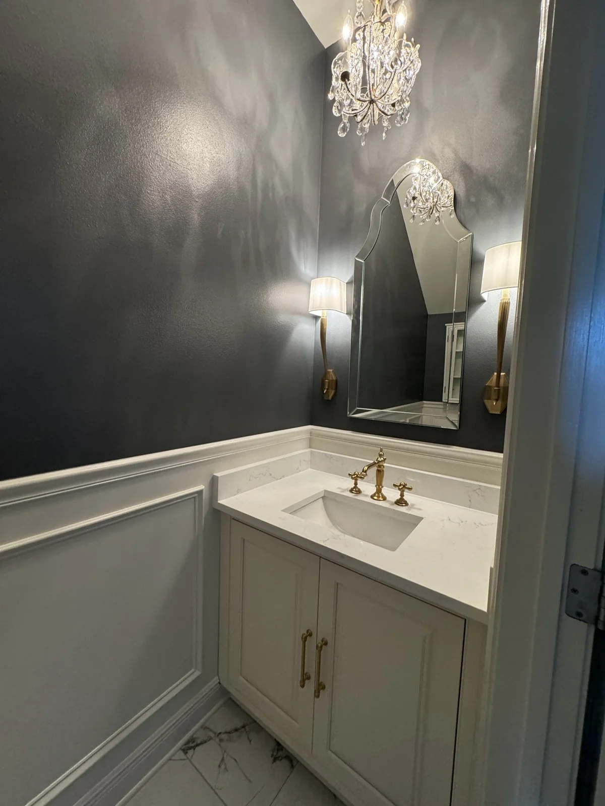Powder room with dark walls, white vanity, brass fixtures and chandelier